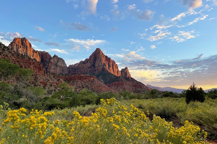A triangular sandstone mountain overlooks green and yellow foliage under a cloudy blue sky.