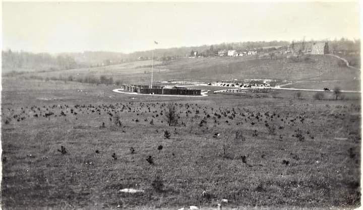 black and white photo of an open field with Fort Necessity in the middle with buildings in the back