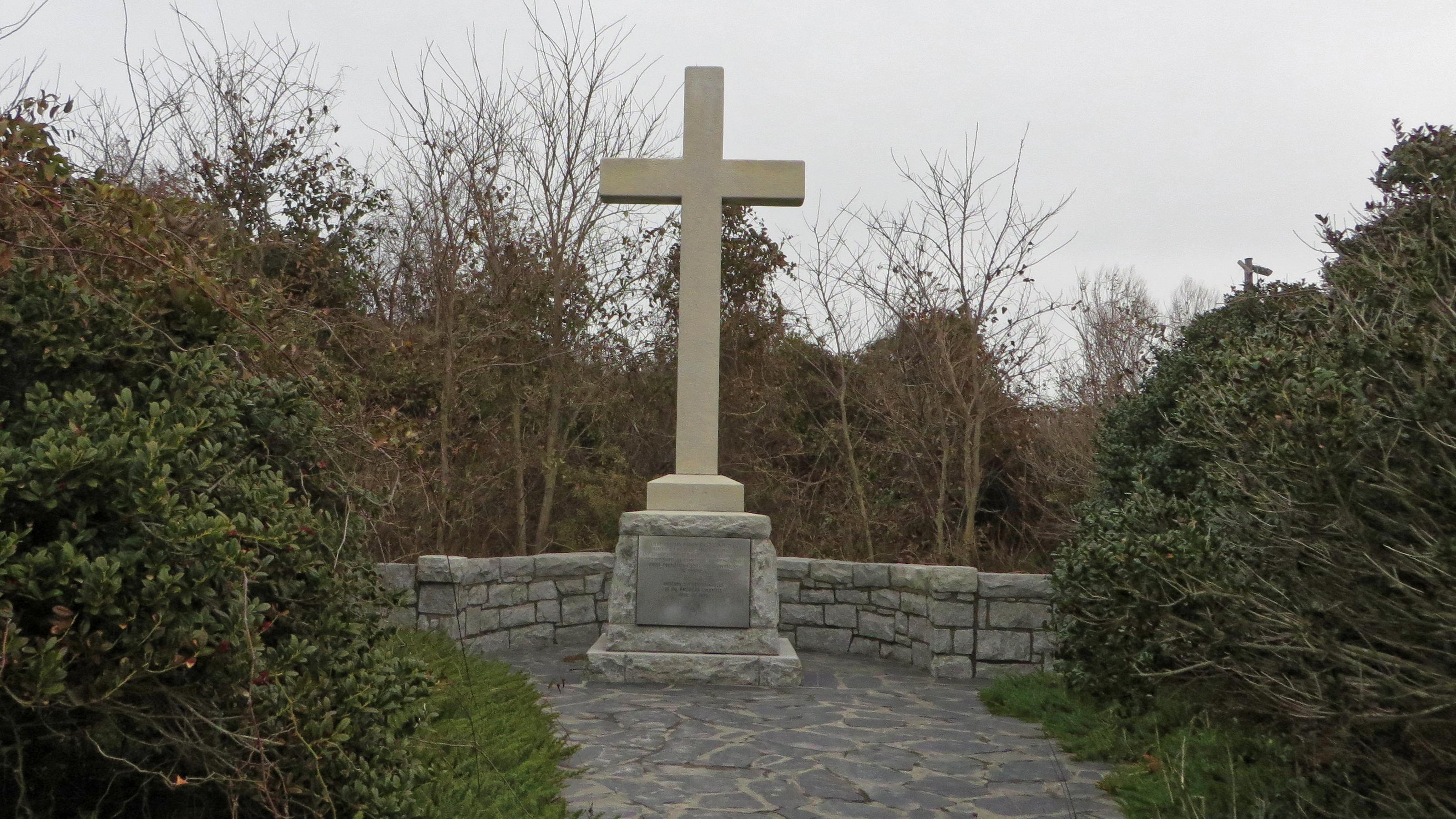 Gray stone cross monument in front of a low stone wall at the end of a flagstone path