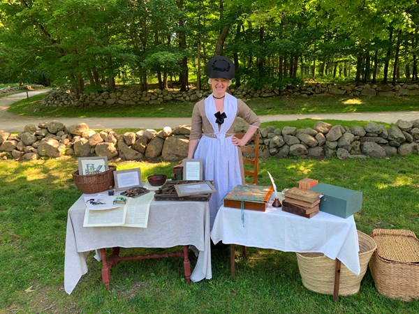 A woman in 1770's clothing stands behind a table filled with materials for printing newspapers.