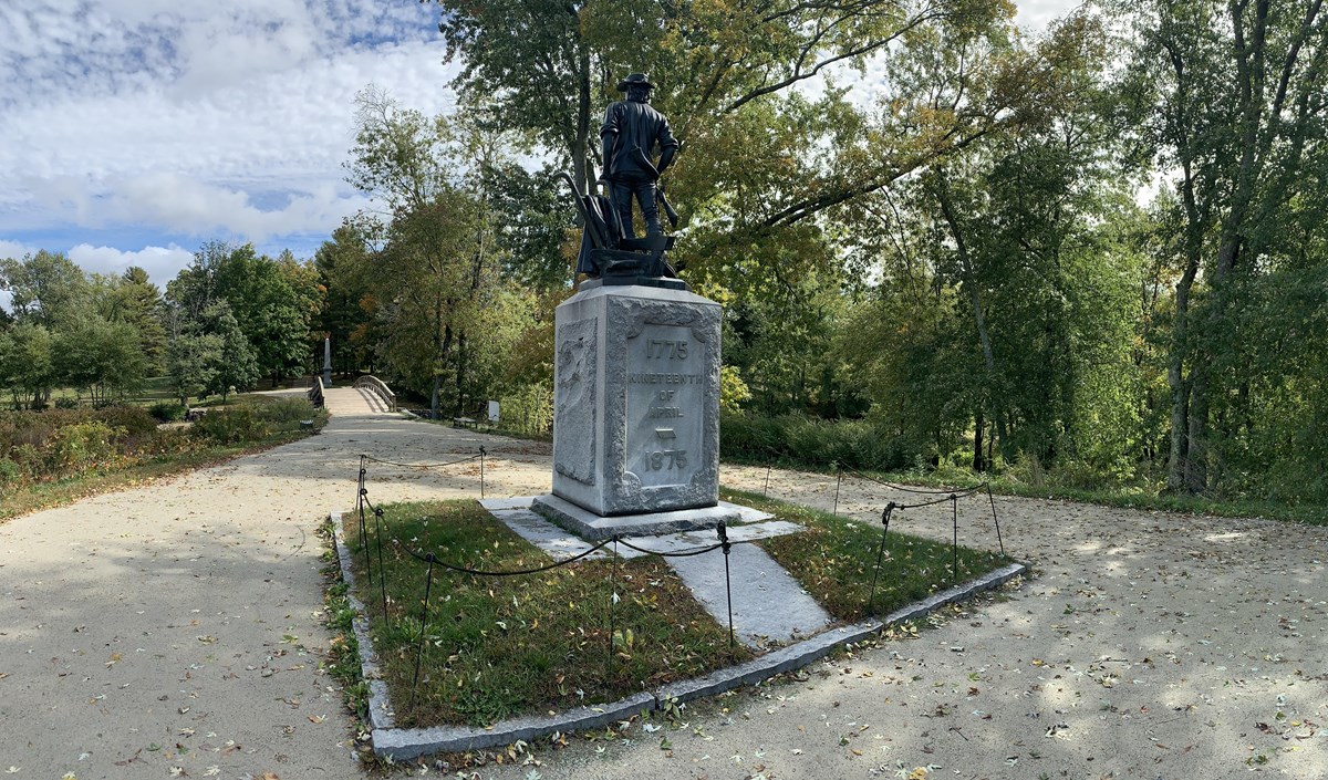 The state of the Minute man stands atop a stone base. The wooden North Bridge is seen in the distanc