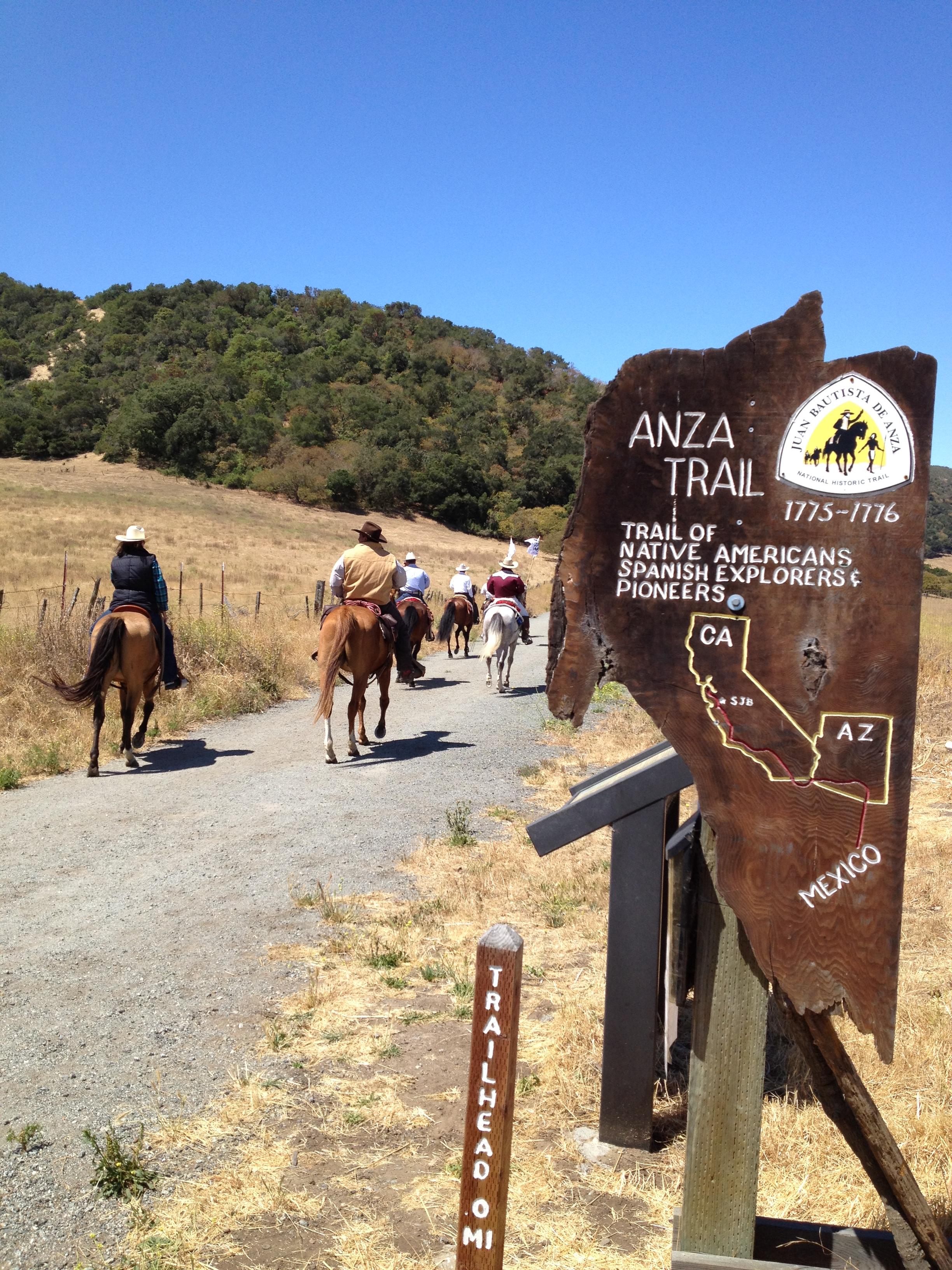 equestrians walk down a dirt trail with an Anza Trail marker