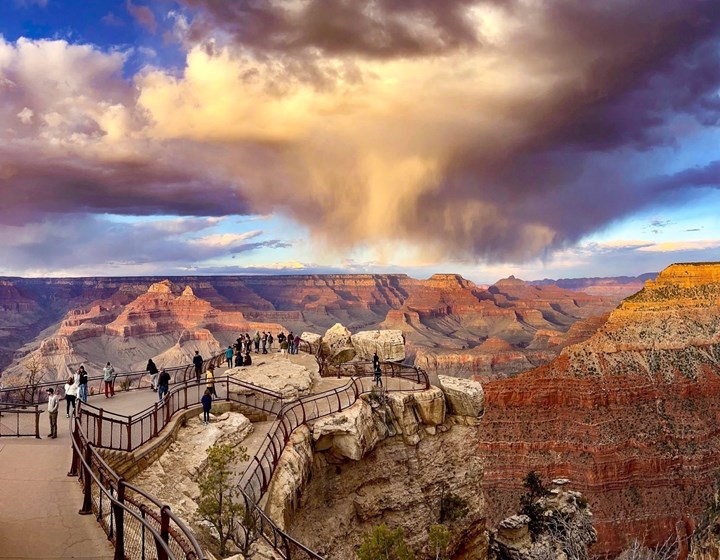At sunset, swirling rain clouds above a vast canyon filled with colorful peaks and cliff walls.