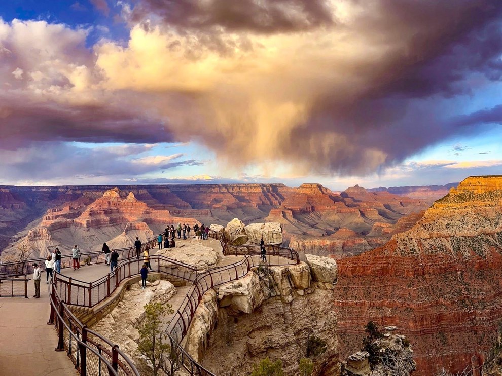 At Sunset, swirling rain clouds above a vast canyon filled with colorful peaks and cliff walls.