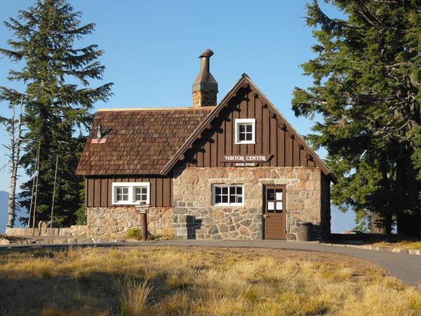 a native stone and wood building, sidewalks through a grassy meadow, towering conifers, and blue sky