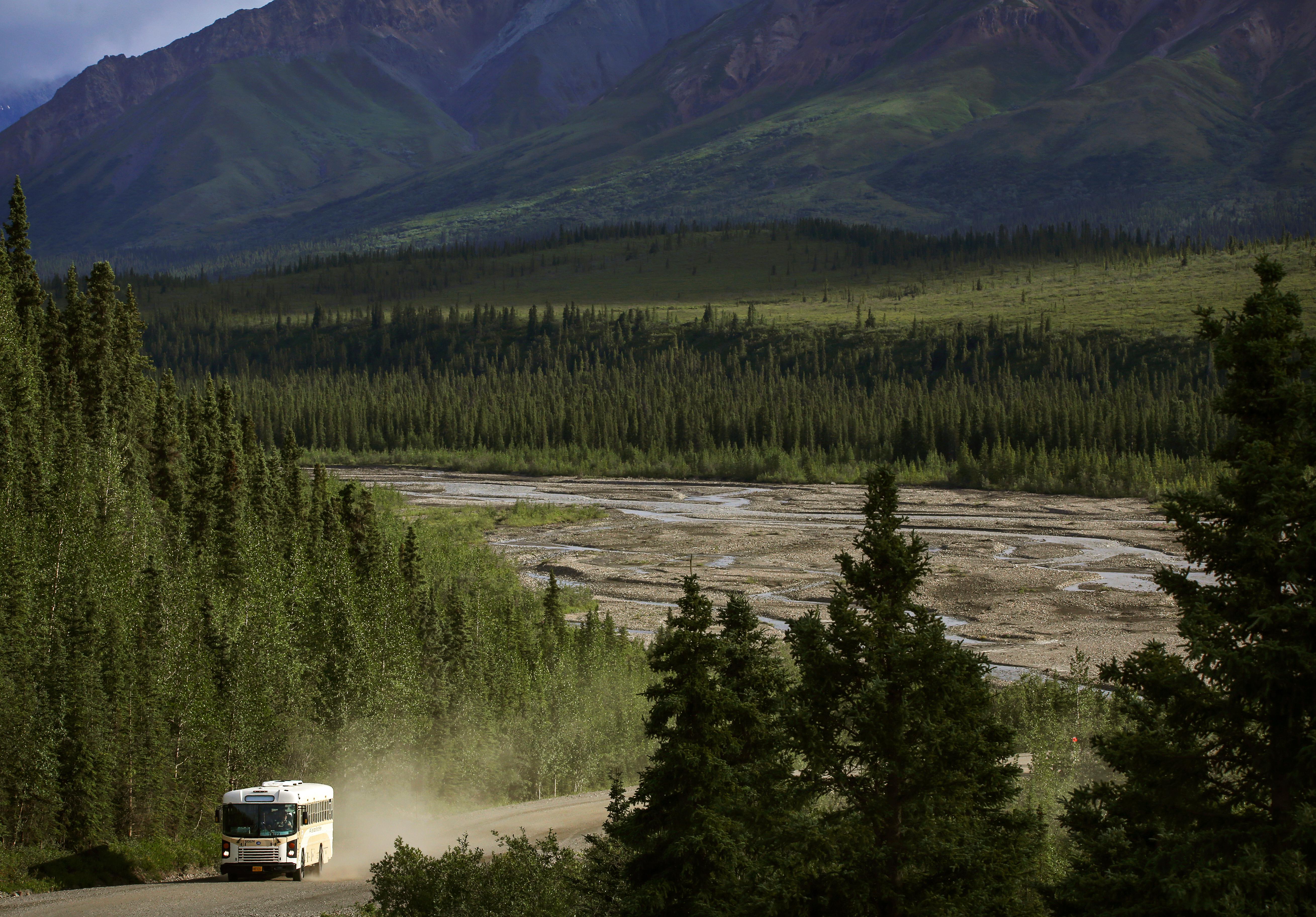 A bus drives along a dusty, gravel road through a forest with mountains and a river in the distance.
