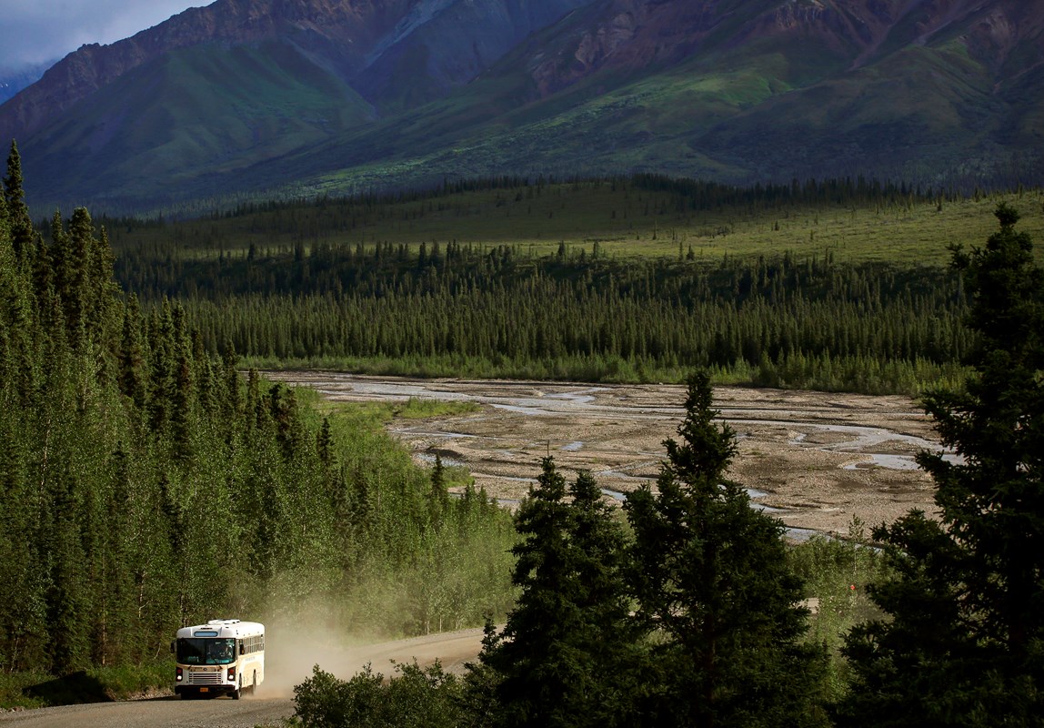 A bus drives along a dusty, gravel road through a forest with mountains and a river in the distance.