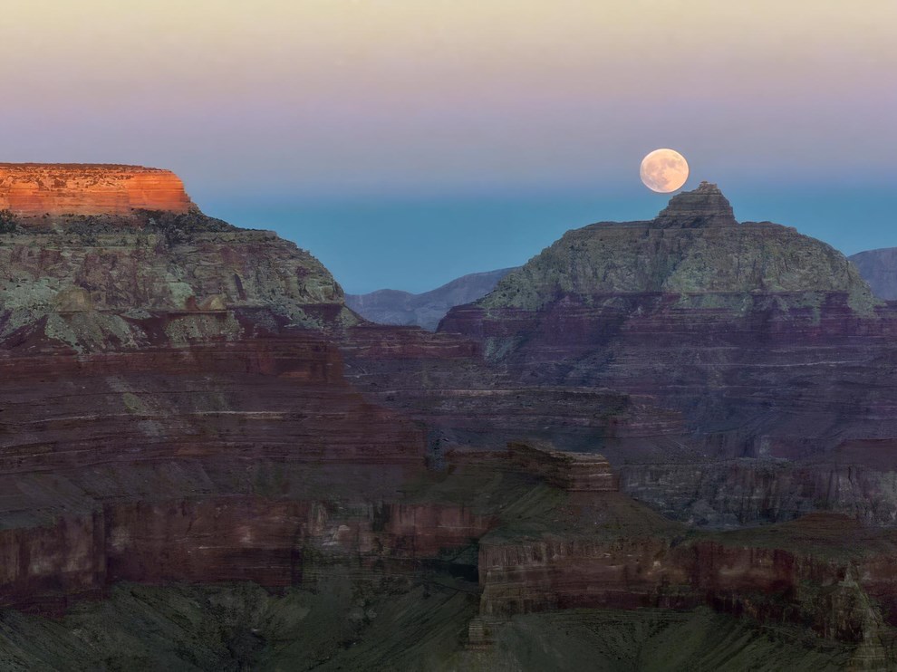 The last glow of sunset across the canyon rim with a full moon rising above a pointed peak
