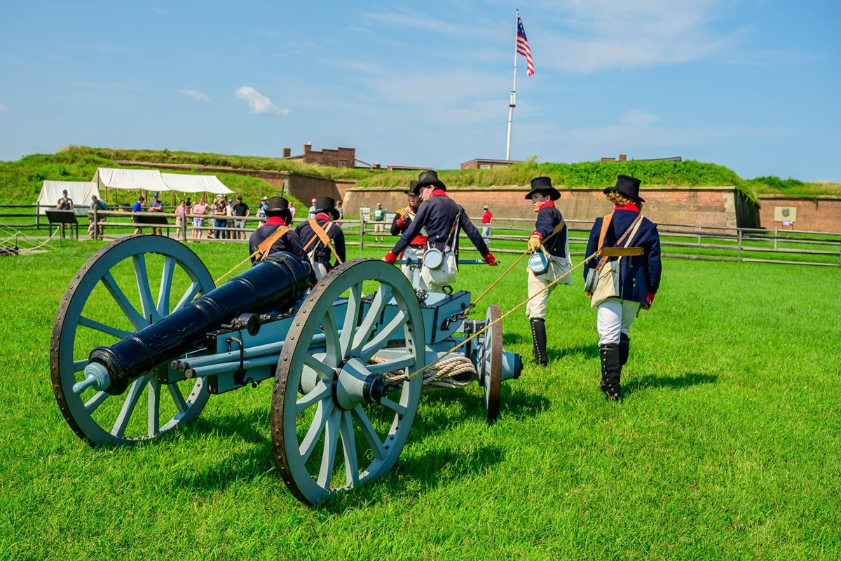 Men in War of 1812 era uniforms pull a cannon over grass outside of a brick fort. 