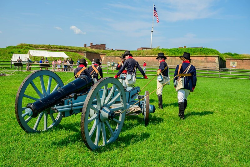 Cannon Demonstration Men in War of 1812 era uniforms pull a cannon over grass outside of a brick fort.