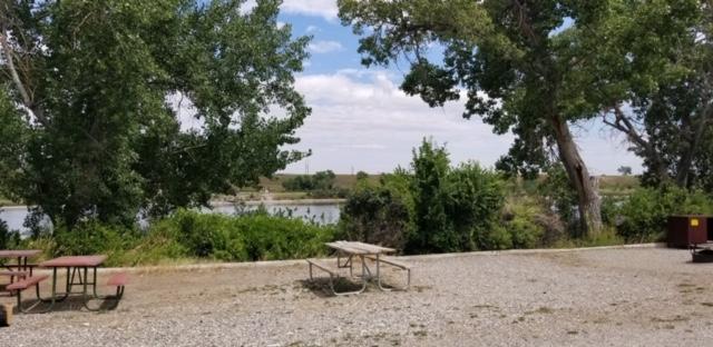 A gravel campground spot with a fire pit and picnic table with trees and afterbay lake in the back.