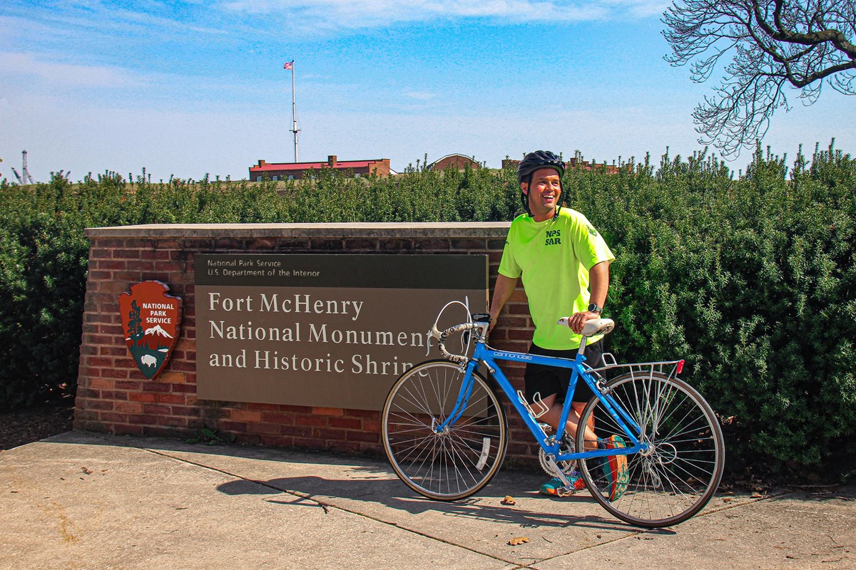 Man in brought yellow shirt with a blue bicycle standing in front of a Fort McHenry identifier sign.
