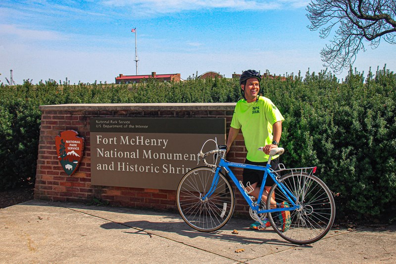 Bike Around Fort McHenry Man in brought yellow shirt with a blue bicycle standing in front of a Fort McHenry identifier sign.