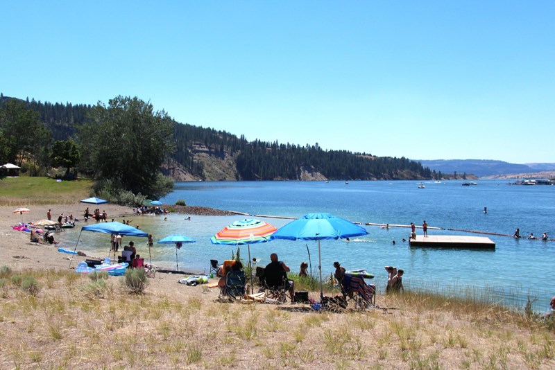 Lake Roosevelt Fort Spokane Day Use Area Scattered beach umbrellas shade various people sitting in chairs at a lakeside beach.