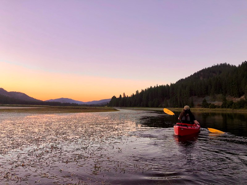 Lake Roosevelt Kettle River Kayaker Warm and purple hues light up the sky and water as a kayaker in a red kayak paddles along.
