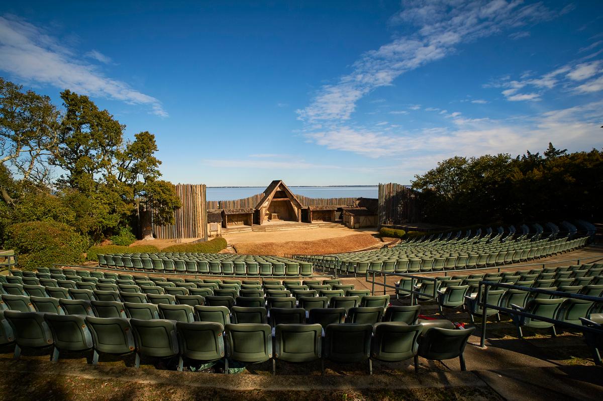 An outdoor theater on the shoreline with green stadium seats, sand stage, and wooden facade