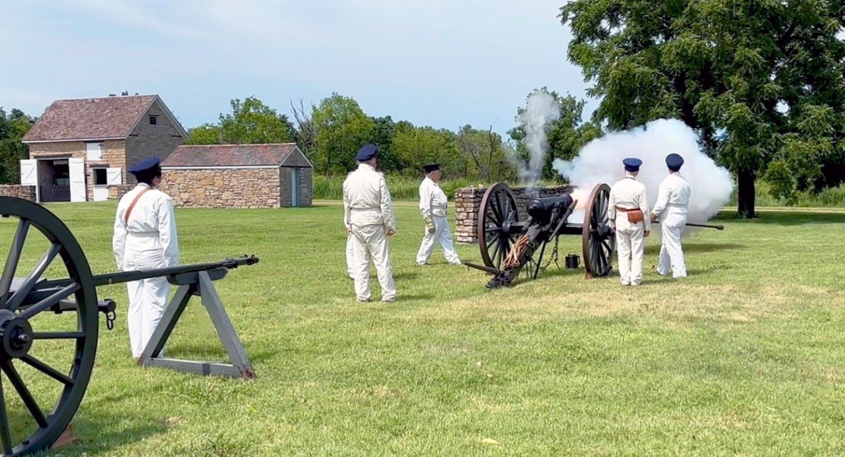 6 reenactors dressed in white are firing a 6lbs field cannon.