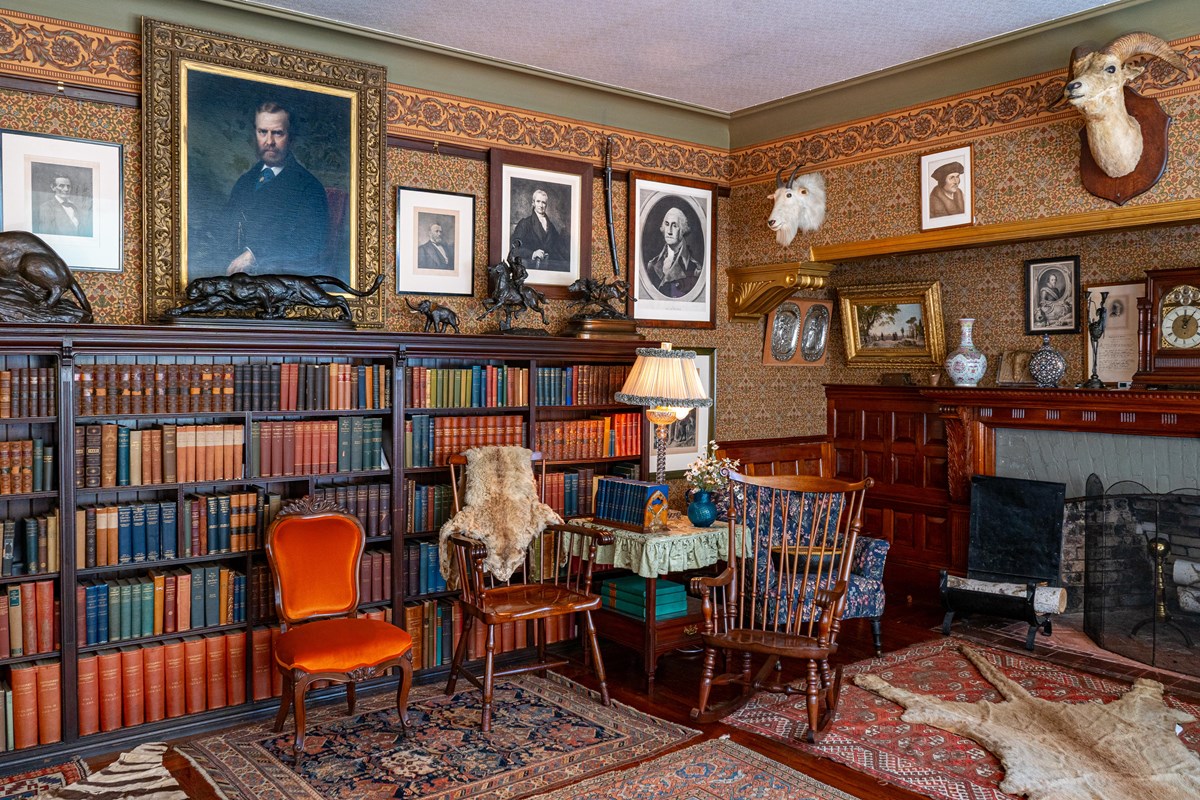 A wallpapered room with bookshelves, chairs, animal trophies, portraits and other mementos.