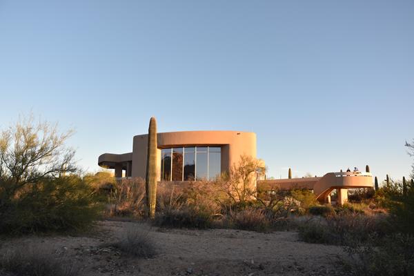 A view of a futuristic looking building perched above a sandy wash, surrounded by saguaros and trees