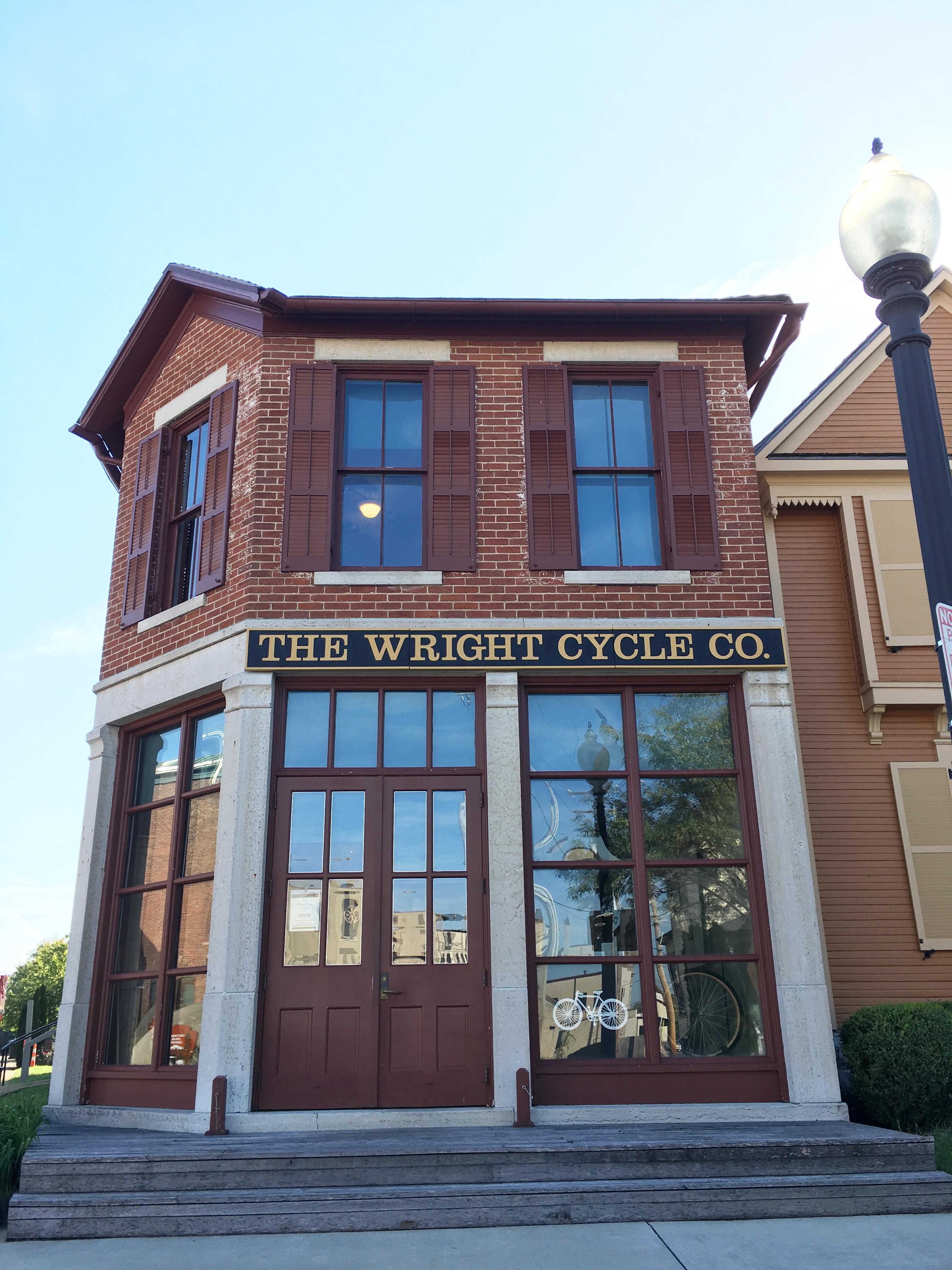 A two-story brick building with tall windows and shutters on them with sign "The Wright Cycle Co."