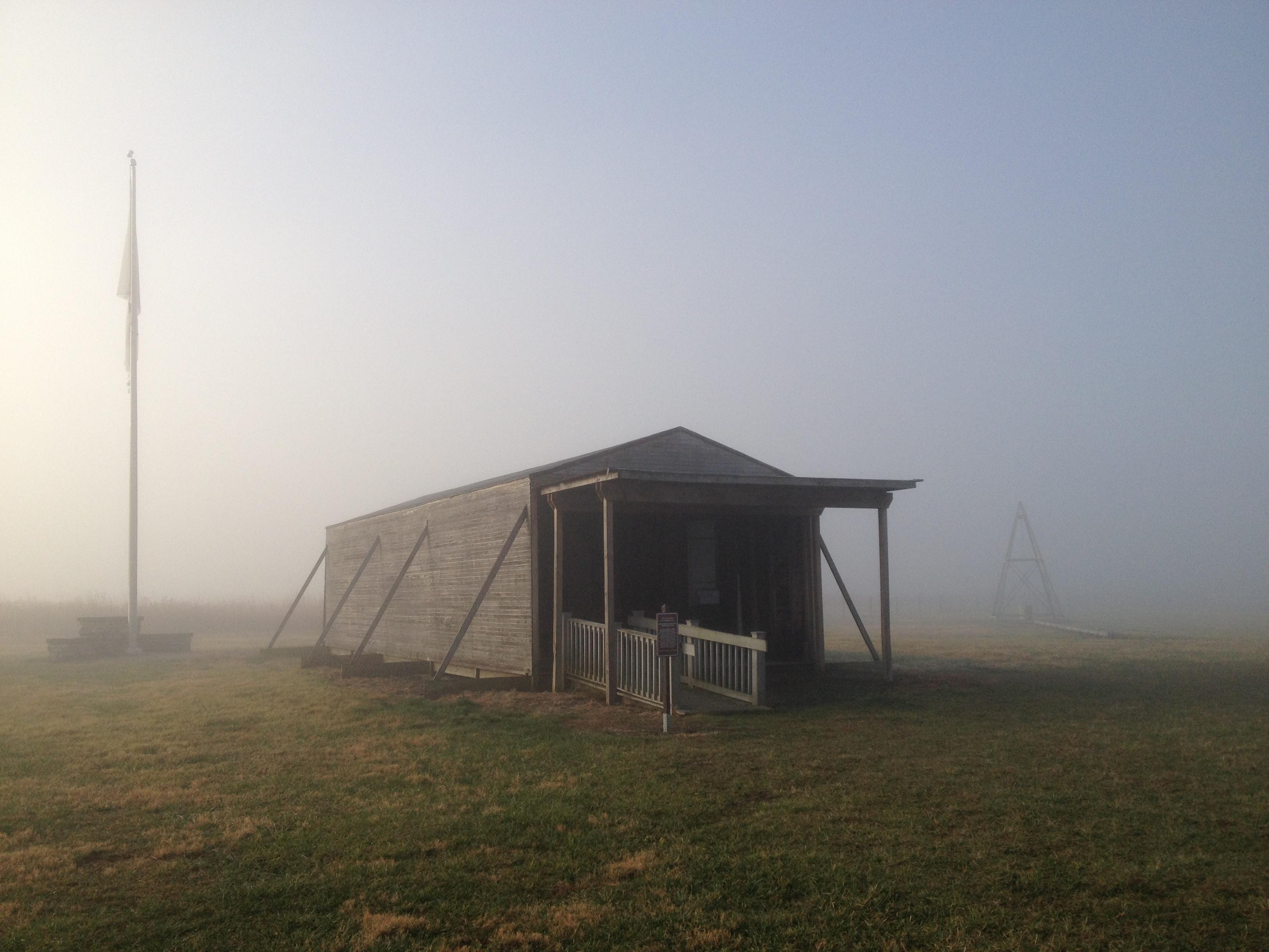 The fog lifts as the suns rays peek through the mist revealing a wood-frame hangar and flag pole.