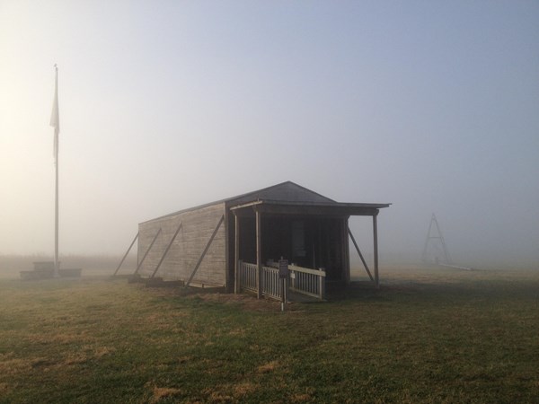 The fog lifts as the suns rays peek through the mist revealing a wood-frame hangar and flag pole.