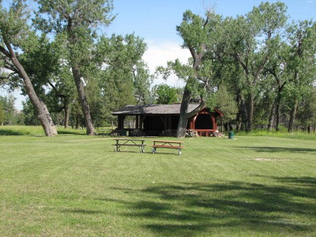A picnic shelter sits in a green lawn with picnic tables and large cottonwood trees.