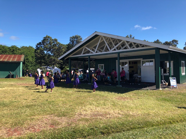 A group of hula dancers in purple clothing gathered outside a green and white wooden building