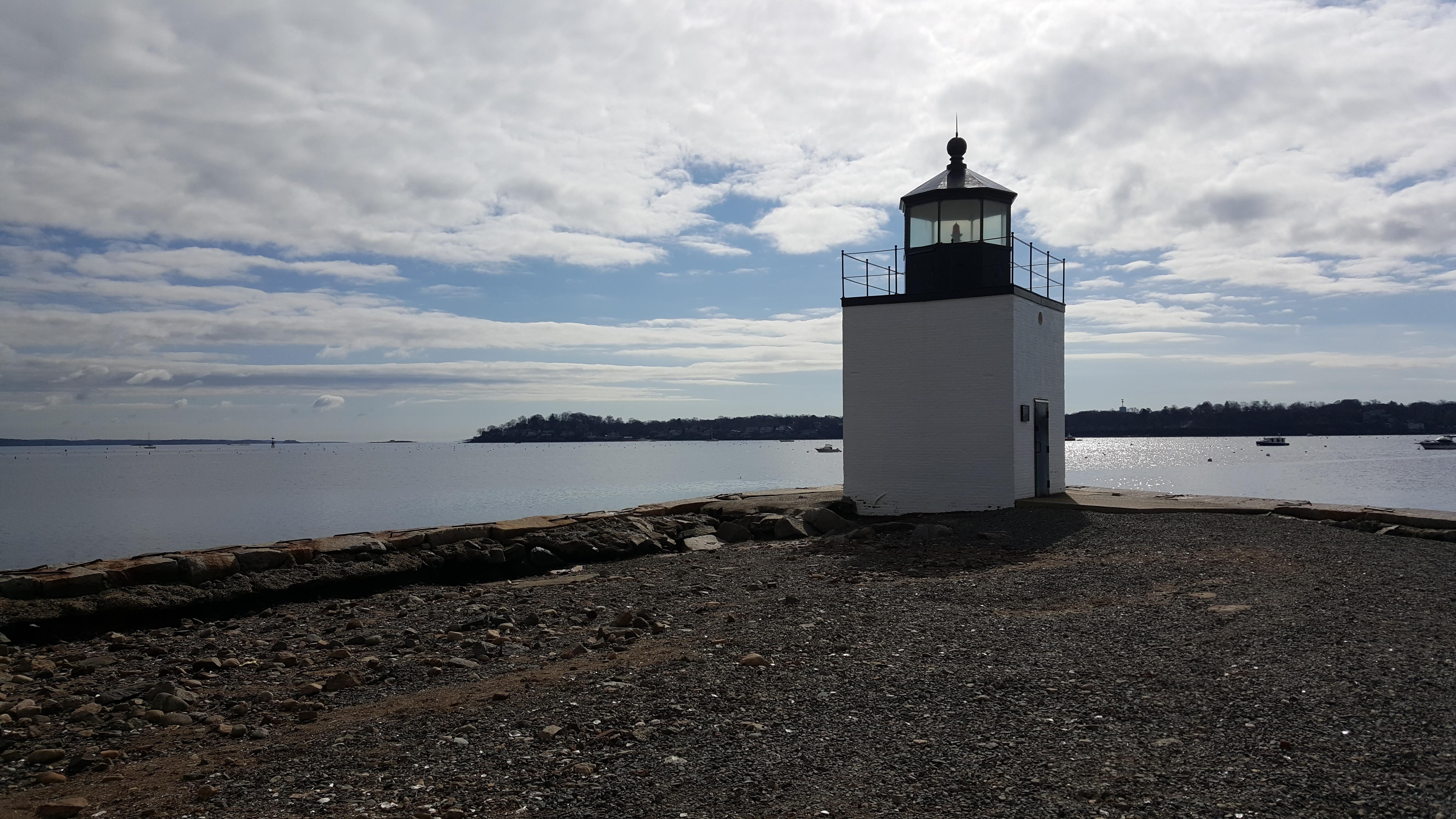 A white rectangular lighthouse approximately 20 feet tall is near the water on a gravel road.