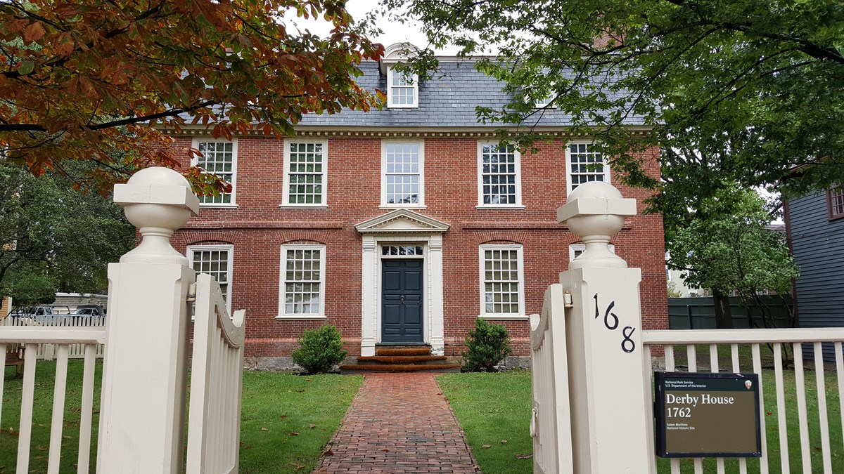 Three story red brick building with white windows and a brick pathway through grasses and trees.
