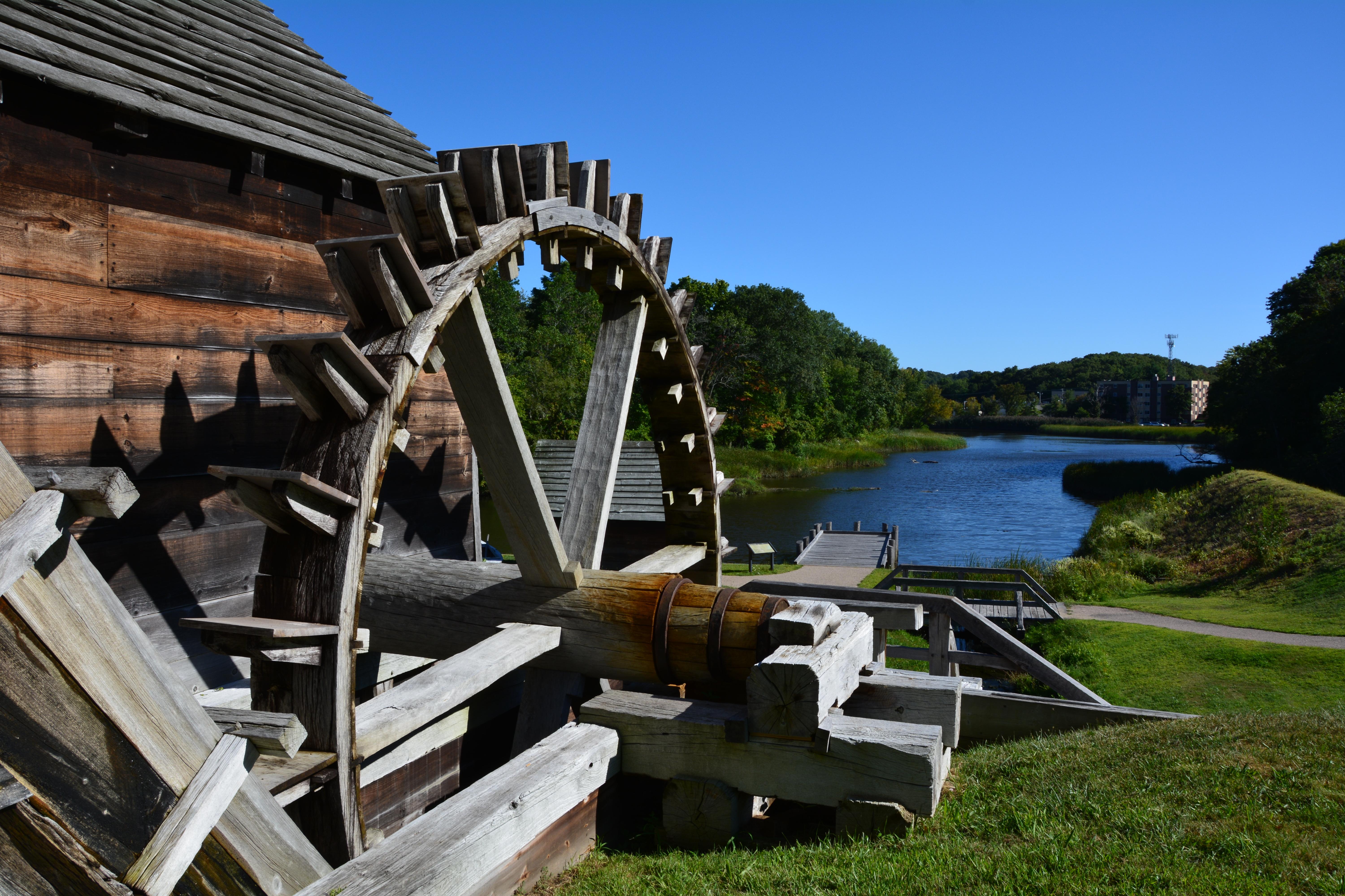Approximately 15' by 15' spoked wooden wheel beside river and under blue sky.