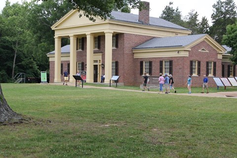 Red-brick visitor center with a yellow columned porch and visitors walking along a path across a lawn