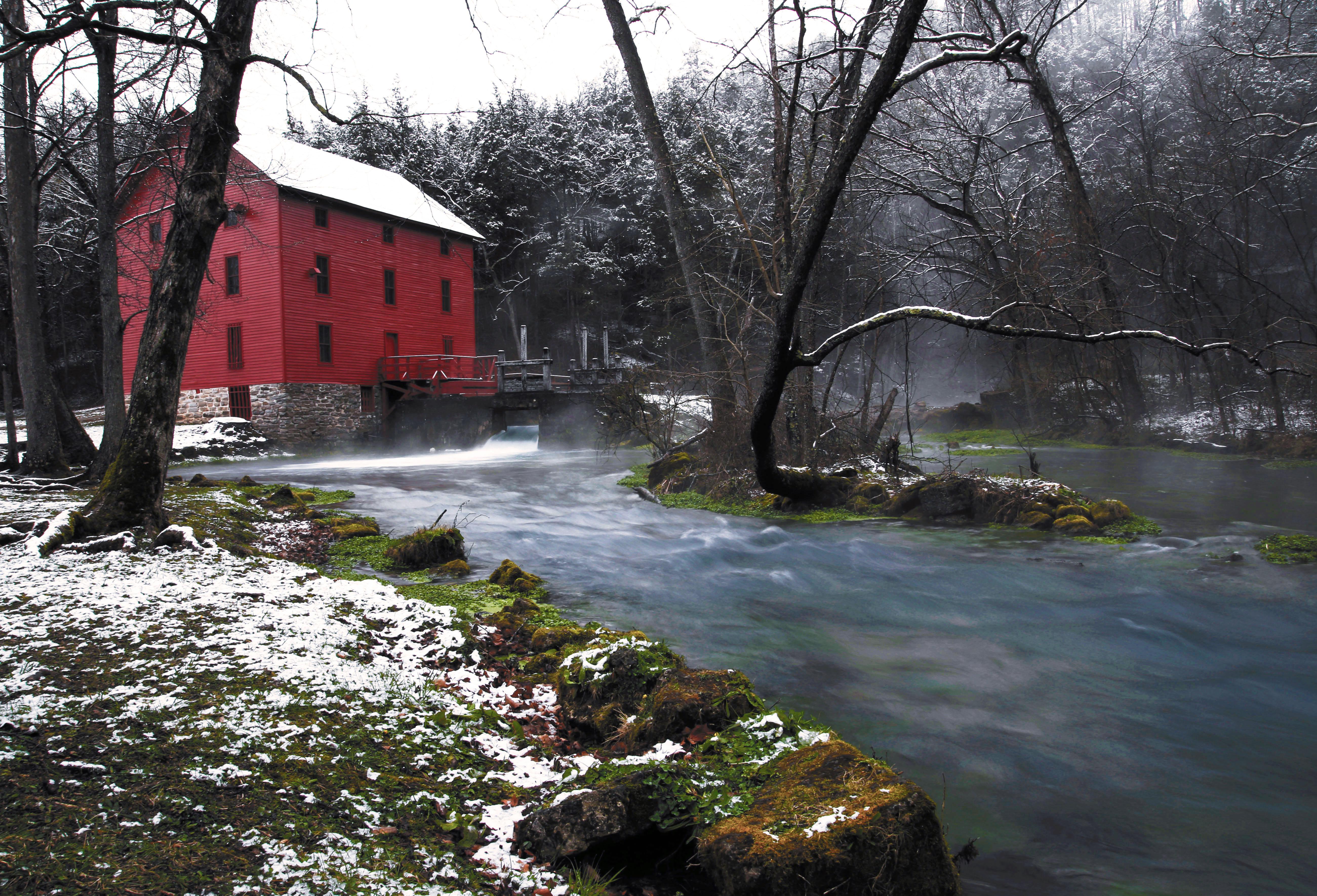 a red mill with snow on roof with a stream emanating from it.