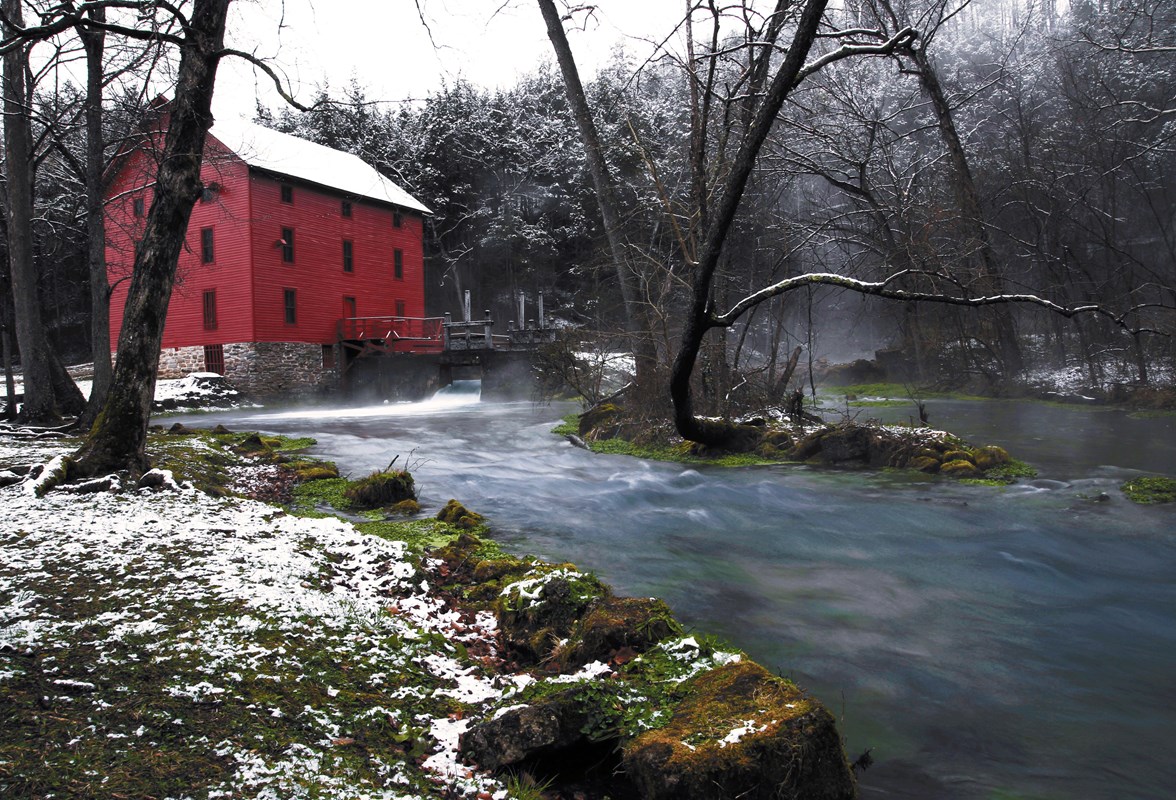 a red mill with snow on roof with a stream emanating from it. 