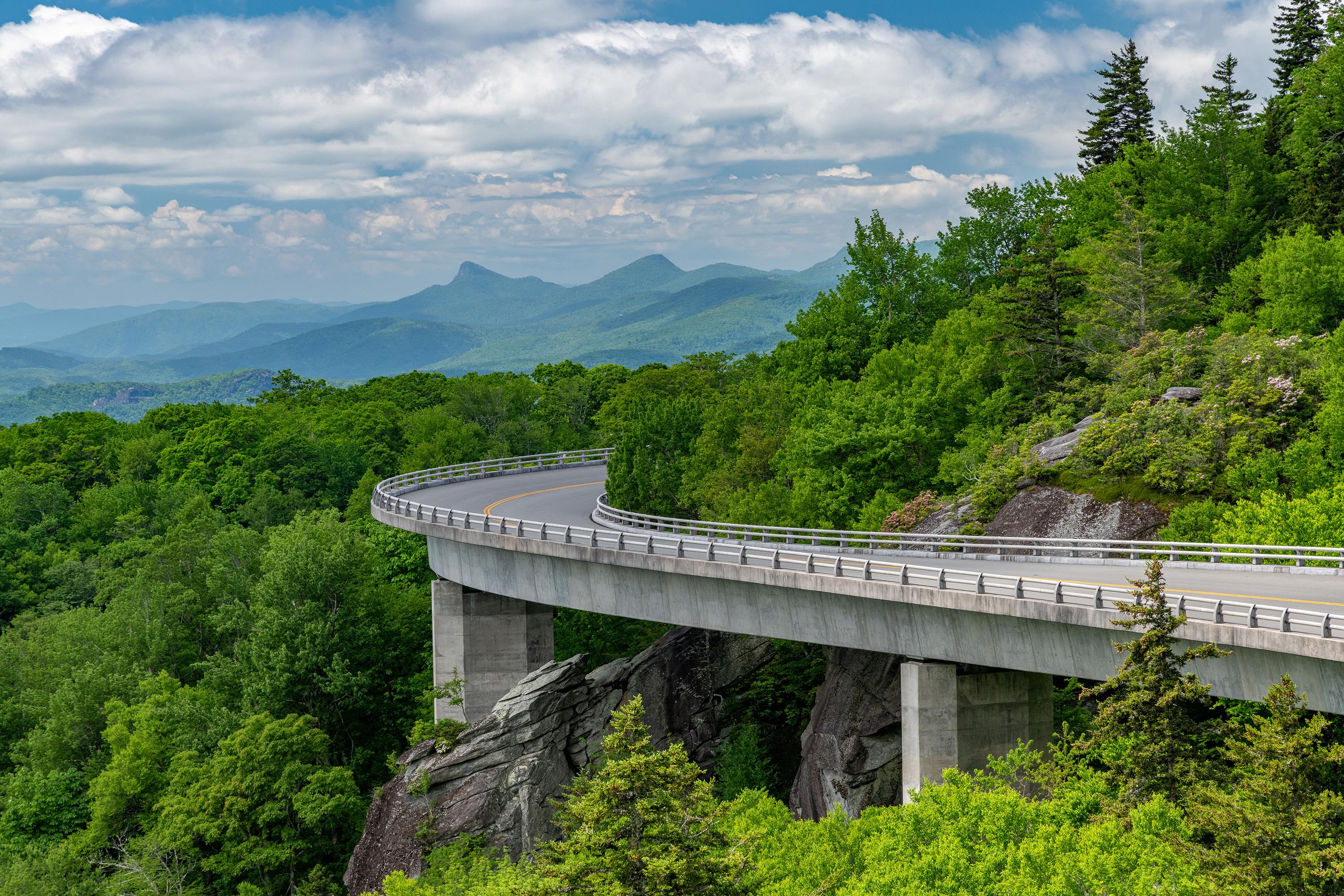 A roadway on piers follows the curve of a mountainside, running toward distant mountains.