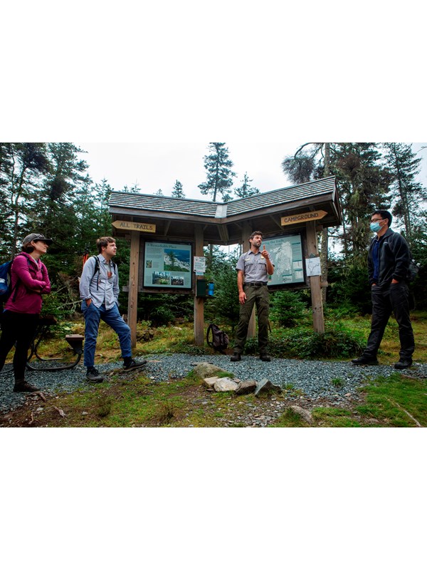 A park ranger and three other adults stand by a wood wayside with area information and directions