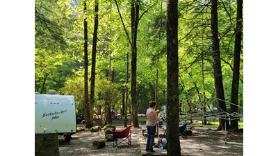 A person standing on a level gravel area setting up a canopy tent, all next to a white trailer.