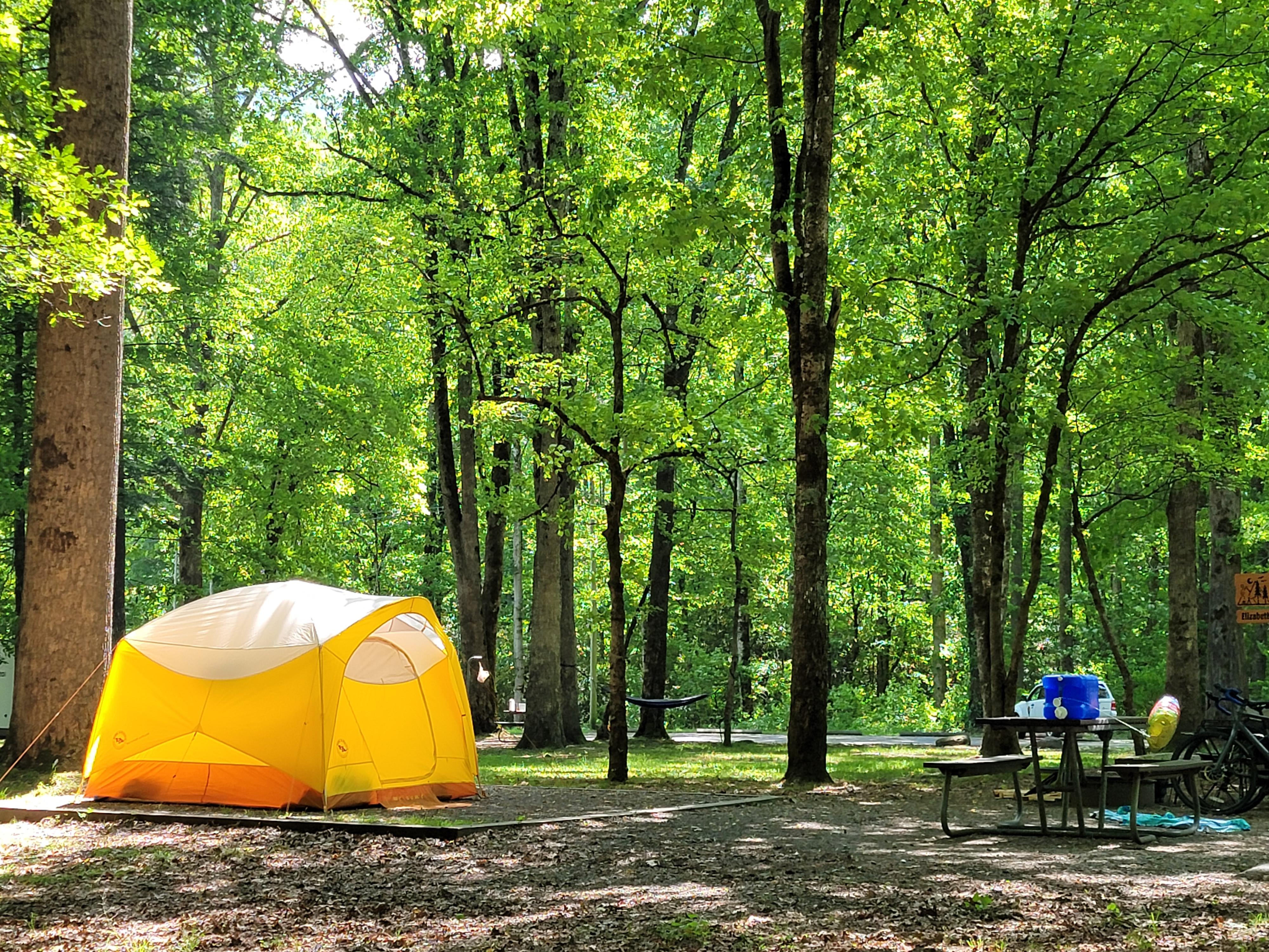 A yellow, orange, and white-colored tent on a level gravel pad near a picnic table and bikes.