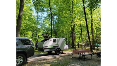 A white trailer and gray SUV parked at a campsite with a red checkered cloth on its picnic table.