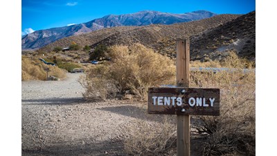 A wooden sign on a wood post reads tents only. Dried bushes extend behind sign right of open area.