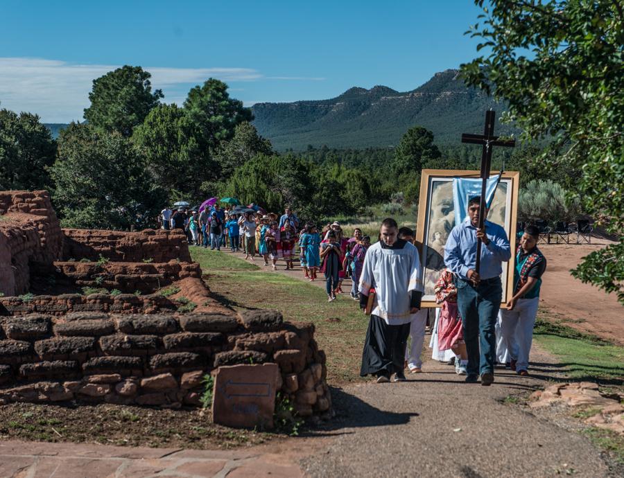 A line of people walk past adobe bricks, trees, and the road.