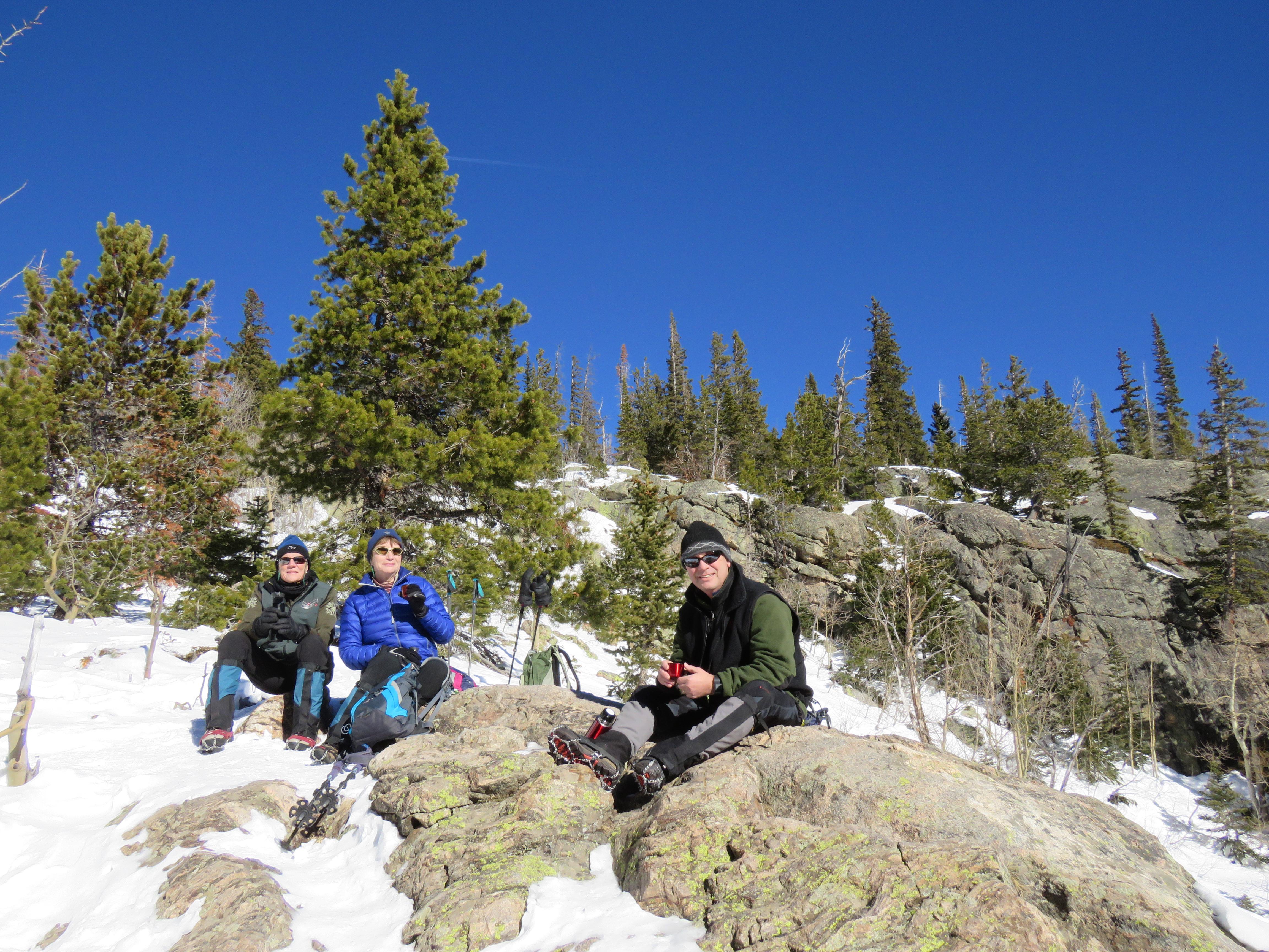 Three people are sitting on rocks on a winter trail. There are patches of snow on the ground.