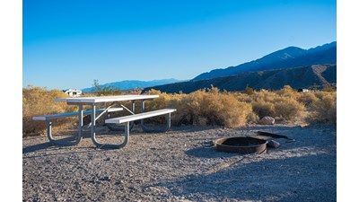 Flat dirt & gravel clearing surrounded by small brown bushes has metal picnic table & fire ring.