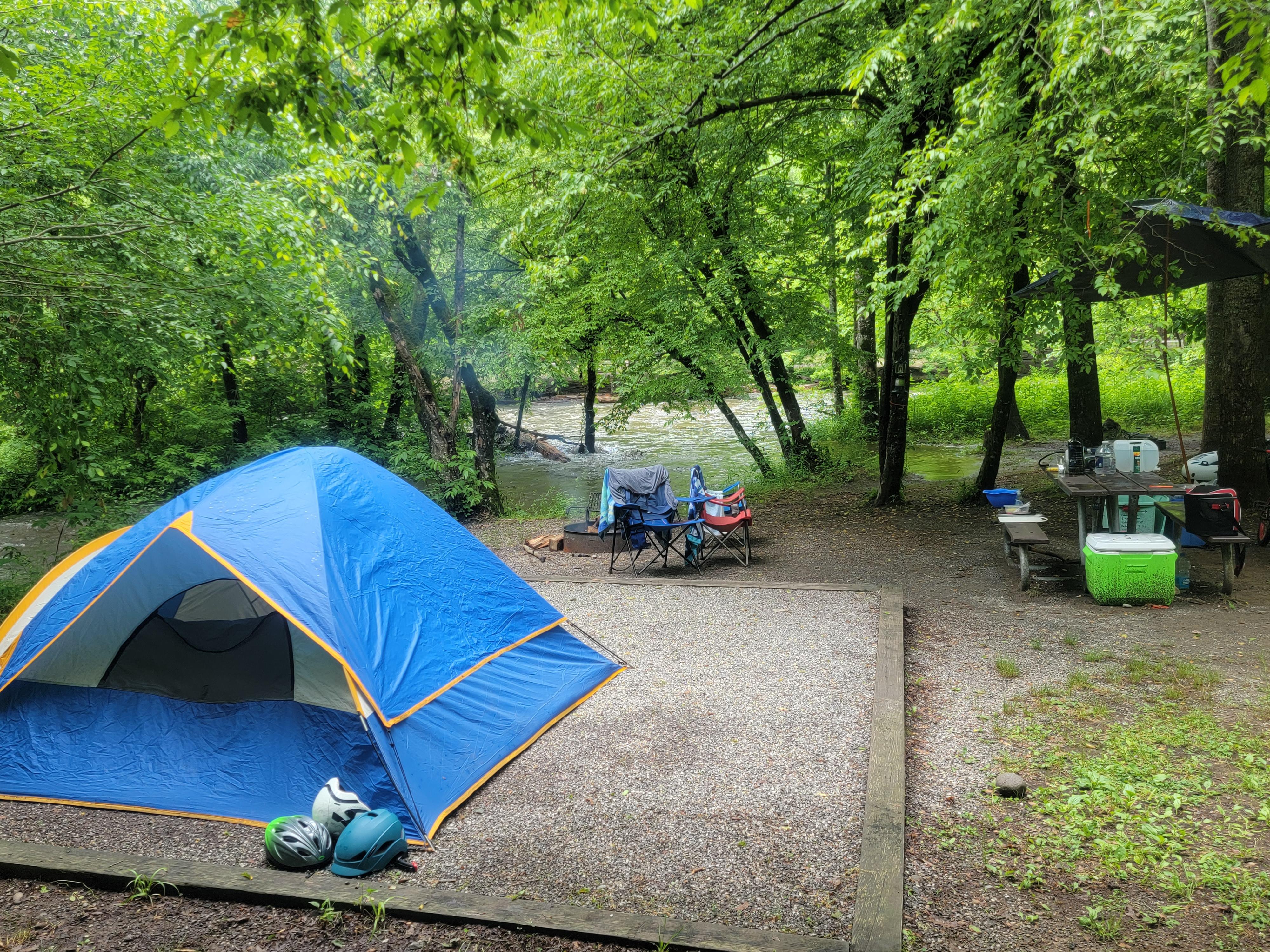 A campsite near flowing water and trees. A blue tent with orange trim sits on the gravel pad.