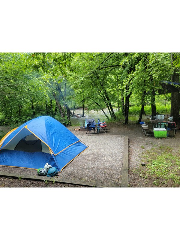 A campsite near flowing water and trees. A blue tent with orange trim sits on the gravel pad.