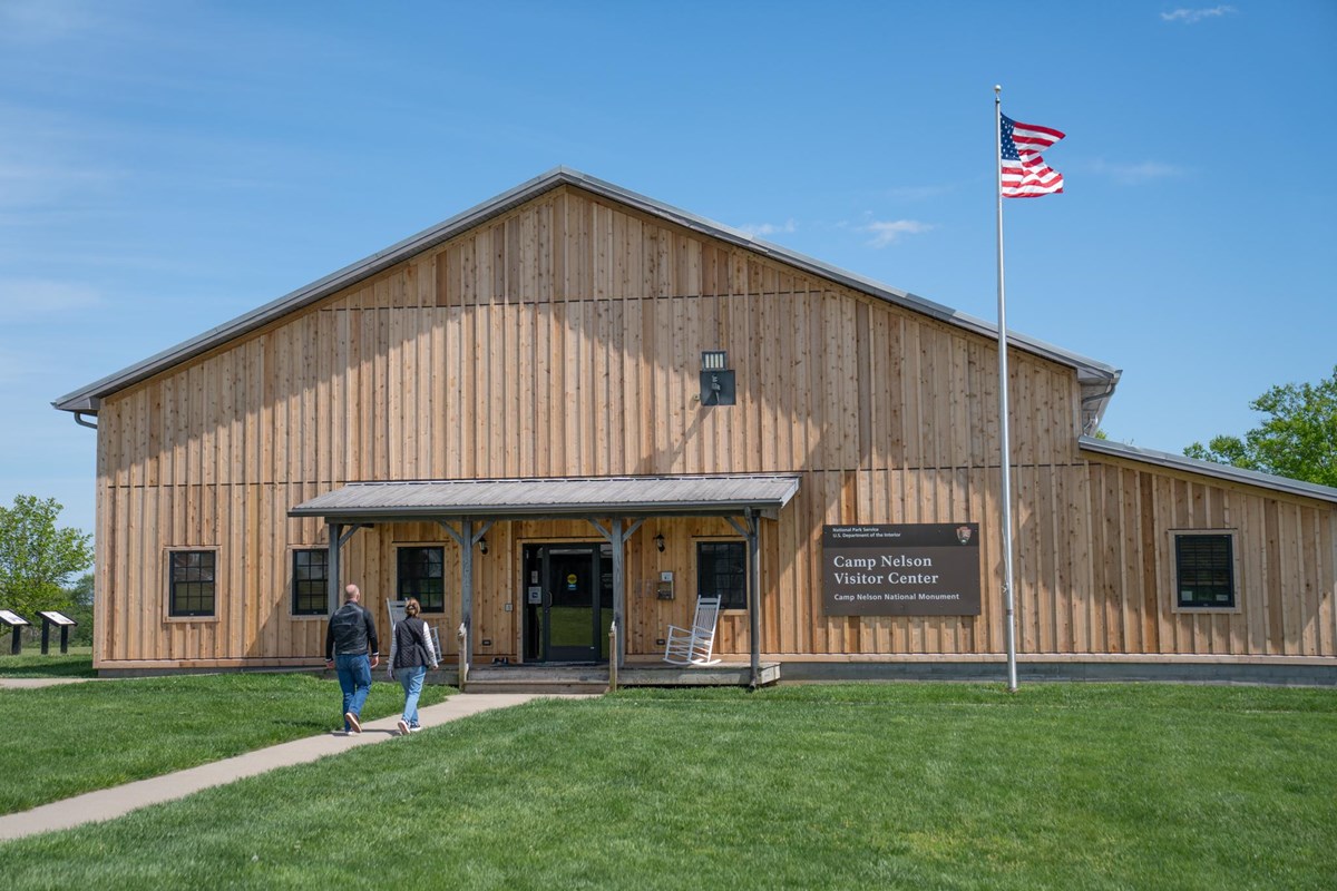 A paved path leads to a gray wooden building with US Flag flying. 