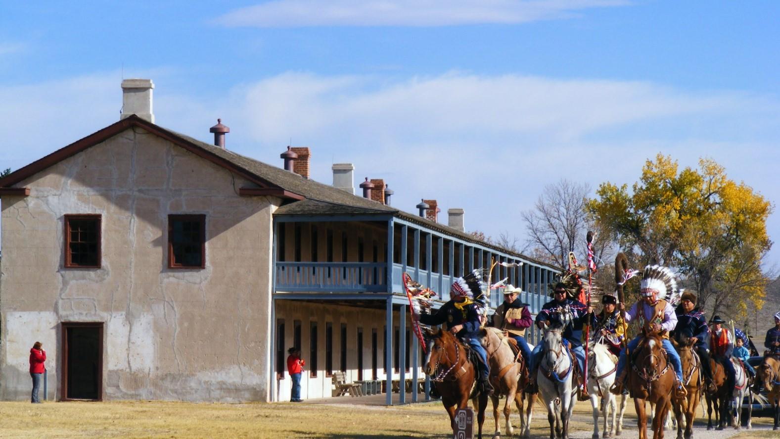 Native American people riding horses - some in traditional regalia - in front the Cavalry Barracks.