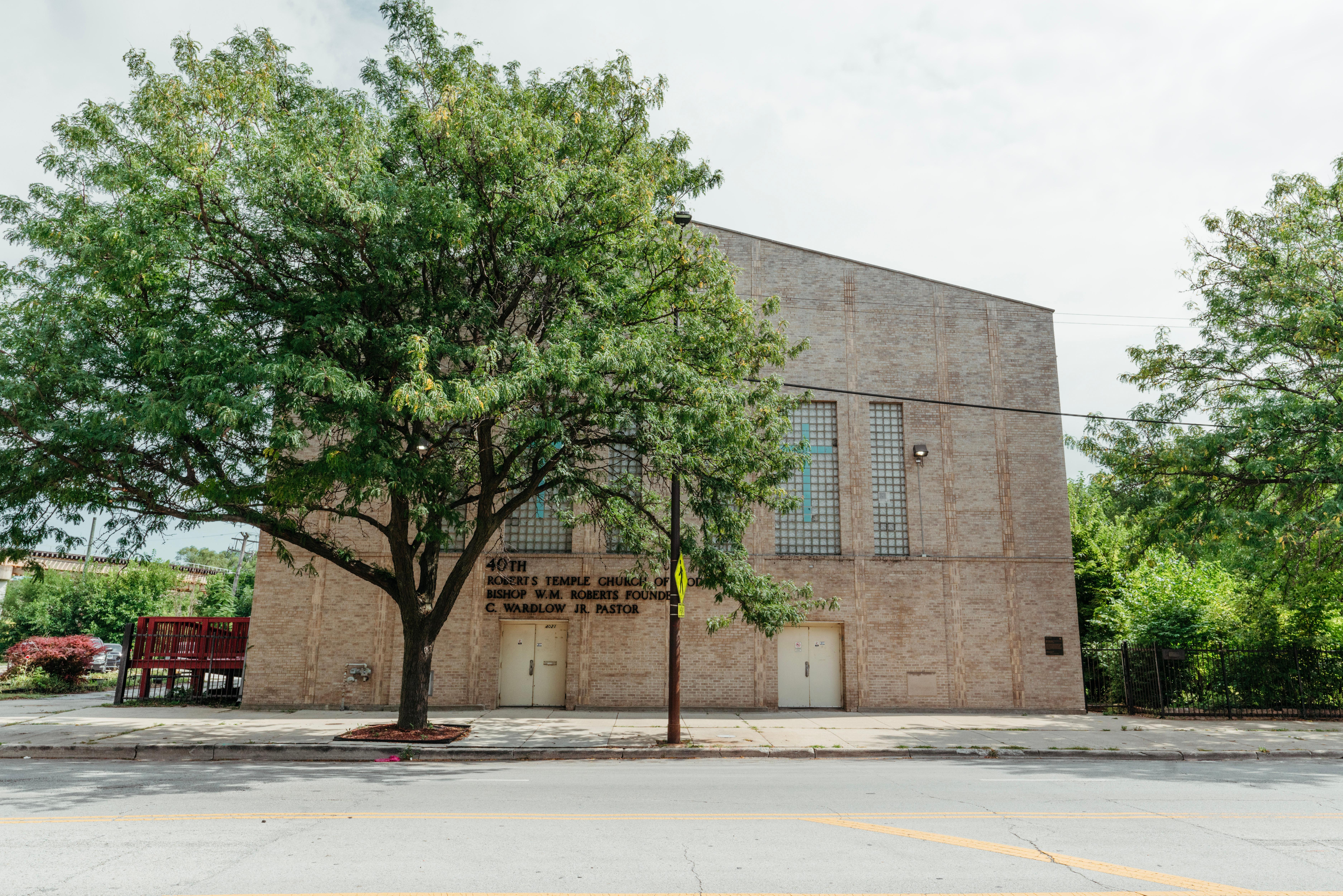 A multi-story tan brick church along a road. A tree stands in front.