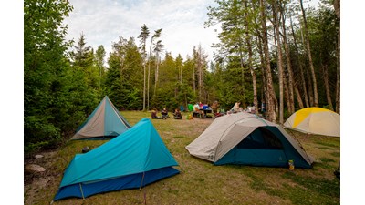 A grassy clearing with four tents and people gathered around picnic tables in the background