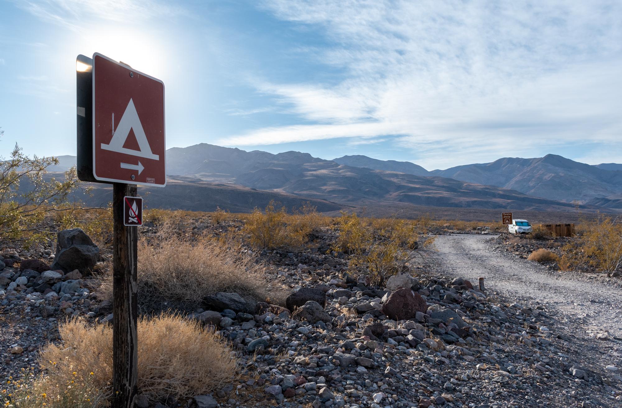 Symbol for tent on a small brown sign on a post stands left of a dirt road. White clouds dot sky.